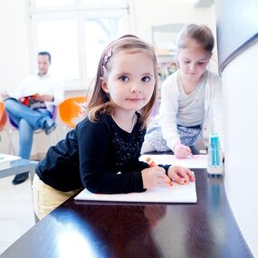 Zwei Kinder malen im Wartezimmer einer Arztpraxis, im Hintergrund sitzt ein Mann.
