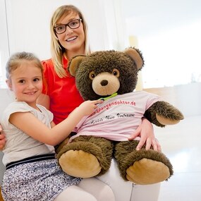 Ein lächelndes Mädchen sitzt mit einer Frau und einem großen Teddybär in einer Zahnarztpraxis.