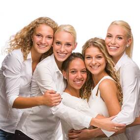 Fünf junge Frauen in weißer Kleidung lächeln gemeinsam vor weißem Hintergrund.